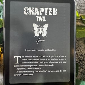 K&ouml;nnte beinhalten: Ein schwarzer Kindle E-Reader, der eine Buchseite mit dem Titel "CHAPTER TWO" und dem Namen "Grace" anzeigt. Eine Schmetterlingsgrafik ist unter dem Titel zentriert. Der Text enth&auml;lt ein Zitat und das Wort "kindle" unten.