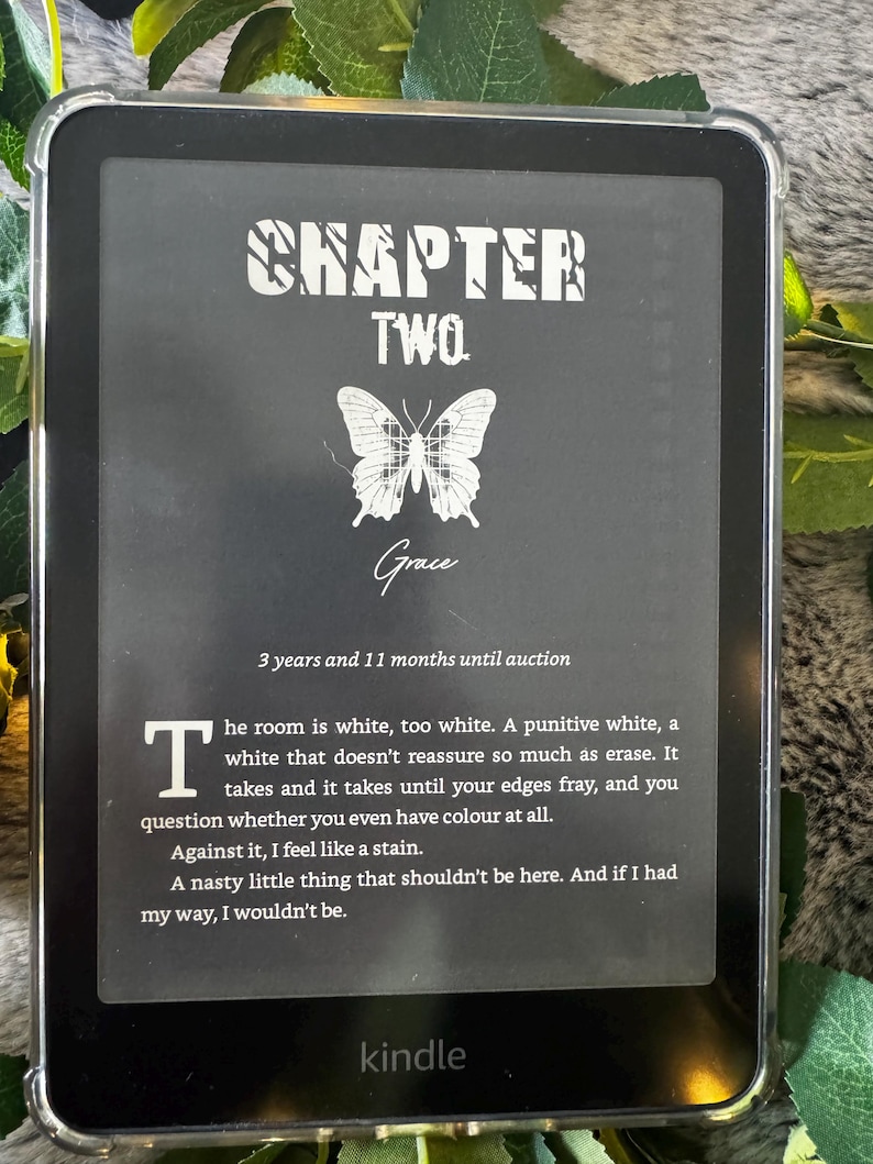 K&ouml;nnte beinhalten: Ein schwarzer Kindle E-Reader, der eine Buchseite mit dem Titel "CHAPTER TWO" und dem Namen "Grace" anzeigt. Eine Schmetterlingsgrafik ist unter dem Titel zentriert. Der Text enth&auml;lt ein Zitat und das Wort "kindle" unten.
