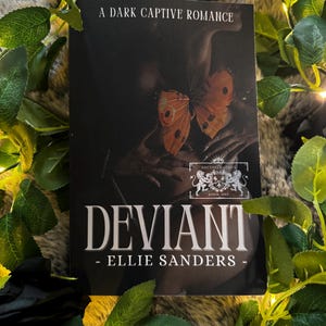 Può includere: Un libro intitolato "Deviant" di Ellie Sanders, con il sottotitolo "A Dark Captive Romance". La copertina presenta una farfalla con ali arancioni e marroni, su sfondo scuro. Il libro è circondato da foglie verdi e luci fatate.