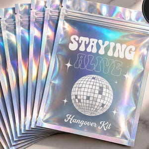 Könnte beinhalten: Ein Set aus zehn holografischen, irisierenden wiederverschließbaren Beuteln mit dem Text STAYING ALIVE und Hangover Kit sowie einer Discokugel-Grafik. Die Beutel haben eine metallisch schimmernde Oberfläche und sind fächerförmig angeordnet.