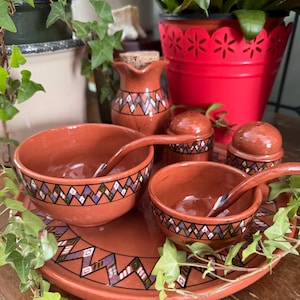 Puede incluir: Un juego de servir de cerámica color terracota con una bandeja redonda grande, dos cuencos con cucharas, una jarrita y dos saleros. Cada pieza está adornada con un patrón geométrico en negro, blanco y morado. El conjunto se exhibe sobre una superficie de madera.