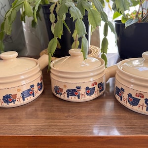 Könnte beinhalten: Drei beige Keramik-Suppenschalen mit Deckel und Griffen. Jede Schale hat einen blauen gepunkteten Rand mit Illustrationen von Hoftieren, darunter Schafe, Kühe und Hühner. Die Schalen sind auf einer Holzoberfläche angeordnet.