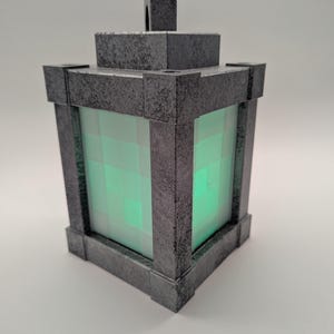 May include: A gray, square lantern with a textured finish. The lantern has a handle on top and a glowing green, pixelated light inside. The frame is a dark gray color.