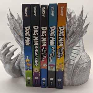 May include: A collection of "Dog Man" graphic novels by Dav Pilkey, including "Dog Man Unleashed" and "Dog Man: Brawl of the Wild." The books are arranged in a row and supported by a silver dragon-shaped bookend.