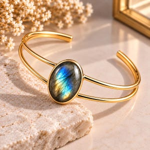 May include: A gold-toned cuff bracelet featuring a central oval-shaped labradorite gemstone. The stone displays iridescent blue and yellow flashes. The bracelet has a double-band design that connects to the bezel setting of the stone.