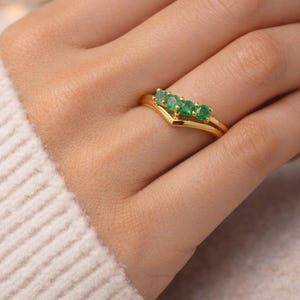 May include: A gold ring with a V-shaped design, set with five emerald-green gemstones. The ring is on a finger, and the hand is wearing a cream-colored sweater. The ring's band is thin and polished.