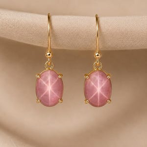 May include: A pair of gold drop earrings featuring oval-shaped pink star stones. Each stone is held by four prongs and displays a distinct six-rayed star pattern. The earrings hang from simple gold hooks with a small spherical bead detail.