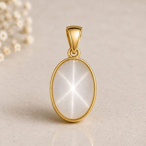 Op de afbeelding: Een gouden hanger met een ovale, witte edelsteen met een stervormig patroon. De hanger heeft een gouden balein om aan een ketting te bevestigen. De edelsteen is gezet in een gouden rand. Ongeveer 2,5 cm lang.