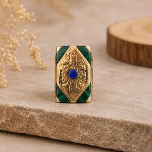 Puede incluir: Un anillo rectangular de oro con una piedra central circular de lapislázuli azul engastada en un intrincado patrón floral en relieve. El diseño está enmarcado por dos paneles triangulares de malaquita verde con bandas oscuras.