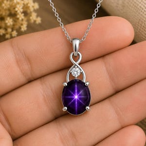 Peut inclure: Un collier pendentif argenté avec un cabochon de saphir étoilé violet profond de forme ovale présentant un effet d'étoile à six branches. La pierre est sertie dans une monture à quatre griffes sous une petite gemme ronde et une boucle infinie.