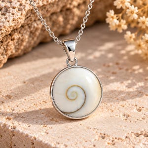 Puede incluir: Un collar con colgante circular que presenta una concha de Shiva blanca pulida con un patrón de espiral marrón natural, engastada en un marco de metal plateado liso y suspendida de una delicada cadena de eslabones plateada.