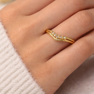 Może przedstawiać: Złoty pierścionek o kształcie litery V. Pierścionek ma małe, przezroczyste kamienie szlachetne wzdłuż górnej krzywizny. Pierścionek jest na palcu, z białym swetrem w tle. To biżuteria.