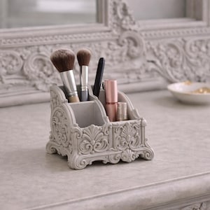 Puede incluir: Un organizador de maquillaje gris claro con detalles ornamentados, que contiene brochas de maquillaje, un bolígrafo y barras de labios. El organizador está sobre una superficie clara, con un espejo y un frasco de perfume en el fondo.