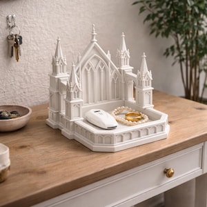 K&ouml;nnte beinhalten: Ein wei&szlig;es Schmucktablett in Form einer Kathedrale mit einem Autoschl&uuml;ssel, einer Perlenkette und einem goldenen Ring. Das Tablett steht auf einer Holzoberfl&auml;che.