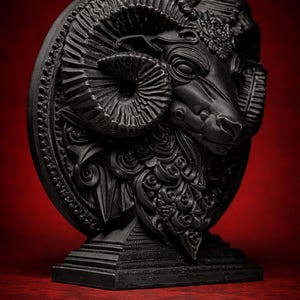 May include: A black ram sculpture with detailed carvings. The ram's head is centered, with large, spiraling horns and intricate facial features. The sculpture is mounted on a pedestal, set against a deep red background.