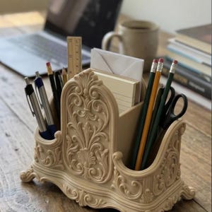 May include: A beige desk organizer featuring intricate scrollwork and floral relief patterns. The structure includes multiple compartments holding various pens, pencils, a wooden ruler, a pair of scissors, and a stack of white note cards.