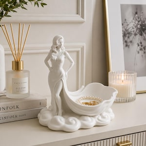 Może przedstawiać: Biała ceramiczna figurka przedstawiająca stojącą postać w długiej sukni, której dół przechodzi w płytką, zakrzywioną miseczkę u podstawy. Postać stoi na chmuropodobnym cokole, a w miseczce znajduje się mały złoty pierścionek oraz perłowa bransoletka.