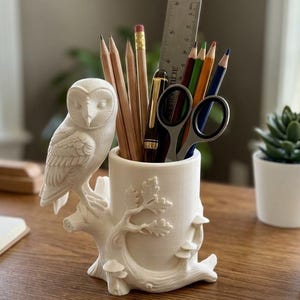 Op de afbeelding: Een witte pennenhouder in de vorm van een uil met een boomstamontwerp. De houder bevat potloden, kleurpotloden, een schaar en een liniaal. De bureau-organizer is gemaakt van keramiek.