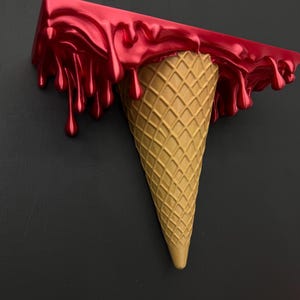 May include: A wall-mounted shelf shaped like an upside-down ice cream cone. The top section is a flat, rectangular surface with a glossy, metallic red finish that appears to be dripping down the sides, while the base is a textured tan cone.
