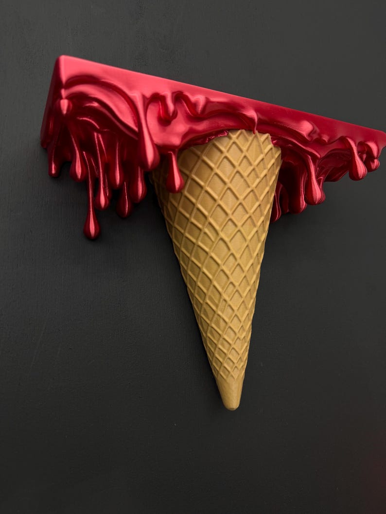 May include: A wall-mounted shelf shaped like an upside-down ice cream cone. The top section is a flat, rectangular surface with a glossy, metallic red finish that appears to be dripping down the sides, while the base is a textured tan cone.