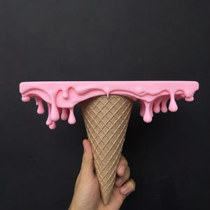 Op de afbeelding: Een plank in de vorm van een omgekeerde ijshoorntje met een beige, getextureerde wafelpatroonbasis en een plat, rechthoekig bovenoppervlak bedekt met dikke, roze, druipende vloeistof die over de randen hangt in ronde, gladde druppels.