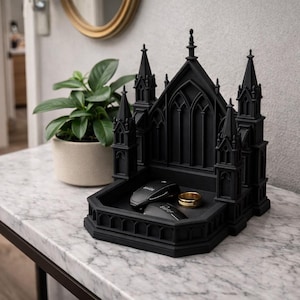K&ouml;nnte beinhalten: Ein schwarzes Tablett in Form einer gotischen Kathedrale enth&auml;lt einen goldenen Ring und zwei schwarze Gegenst&auml;nde. Das Tablett steht auf einem Tisch mit Marmorplatte. Eine brennende Kerze und eine Topfpflanze sind im Hintergrund.