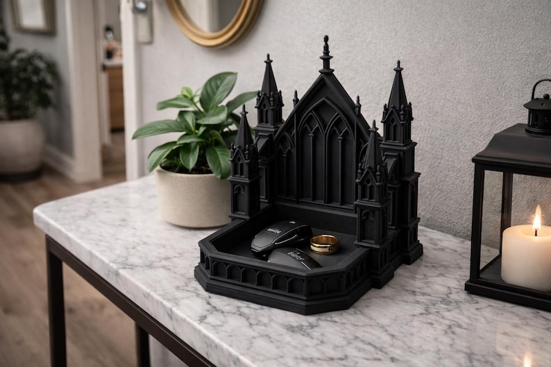 K&ouml;nnte beinhalten: Ein schwarzes Tablett in Form einer gotischen Kathedrale enth&auml;lt einen goldenen Ring und zwei schwarze Gegenst&auml;nde. Das Tablett steht auf einem Tisch mit Marmorplatte. Eine brennende Kerze und eine Topfpflanze sind im Hintergrund.