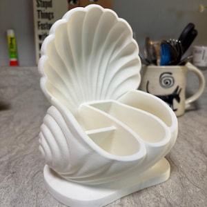 May include: A white seashell-shaped desk organizer with three compartments. The organizer has a textured, ridged design and sits on a flat base. The background includes a mug and a vintage-style sign.