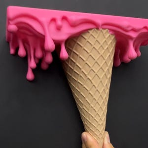 May include: A wall shelf shaped like an upside-down ice cream cone with a tan waffle-textured cone base and a bright pink rectangular top section featuring a realistic dripping liquid effect that hangs down over the edges of the shelf surface.