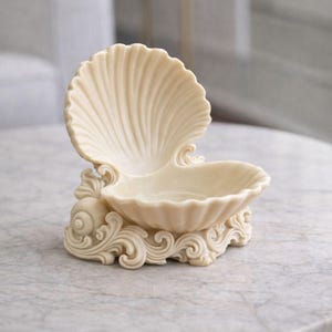 Op de afbeelding: Crèmekleurige, schelpvormige decoratieve schaal. De schaal heeft een groot, open schelpontwerp met een geschulpte rand, ondersteund door een basis met sierlijke, wervelende details. De schaal is gemaakt van een glad, stevig materiaal.