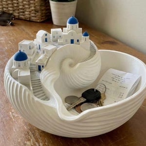 May include: A white decorative bowl with a seashell design, featuring a miniature Santorini village with blue-domed roofs. The bowl contains keys, coins, and a receipt, ideal for holding small items.