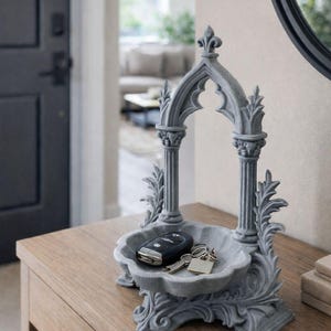 May include: A gray, gothic-style key holder with a decorative arch and ornate detailing. The tray holds a car key and a set of keys. The holder sits on a wooden table, showcasing a blend of functionality and aesthetic appeal.