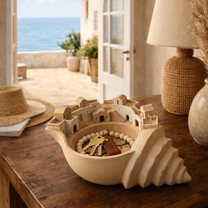 May include: A decorative seashell-shaped bowl made of light beige ceramic. The bowl is filled with keys, coins, and a beaded necklace. A miniature village scene is sculpted along the rim. The bowl sits on a dark wooden table.