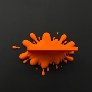 May include: An orange, splash-shaped wall shelf against a dark gray background. The shelf is a solid, horizontal bar integrated into the splash design. The shelf is a vibrant orange color, contrasting with the dark backdrop.