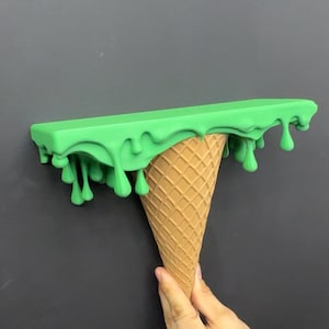 May include: A wall-mounted shelf featuring a flat, rectangular top surface in bright green with a realistic dripping effect along the edges, supported by a textured, tan-colored cone base that resembles a classic waffle ice cream cone.