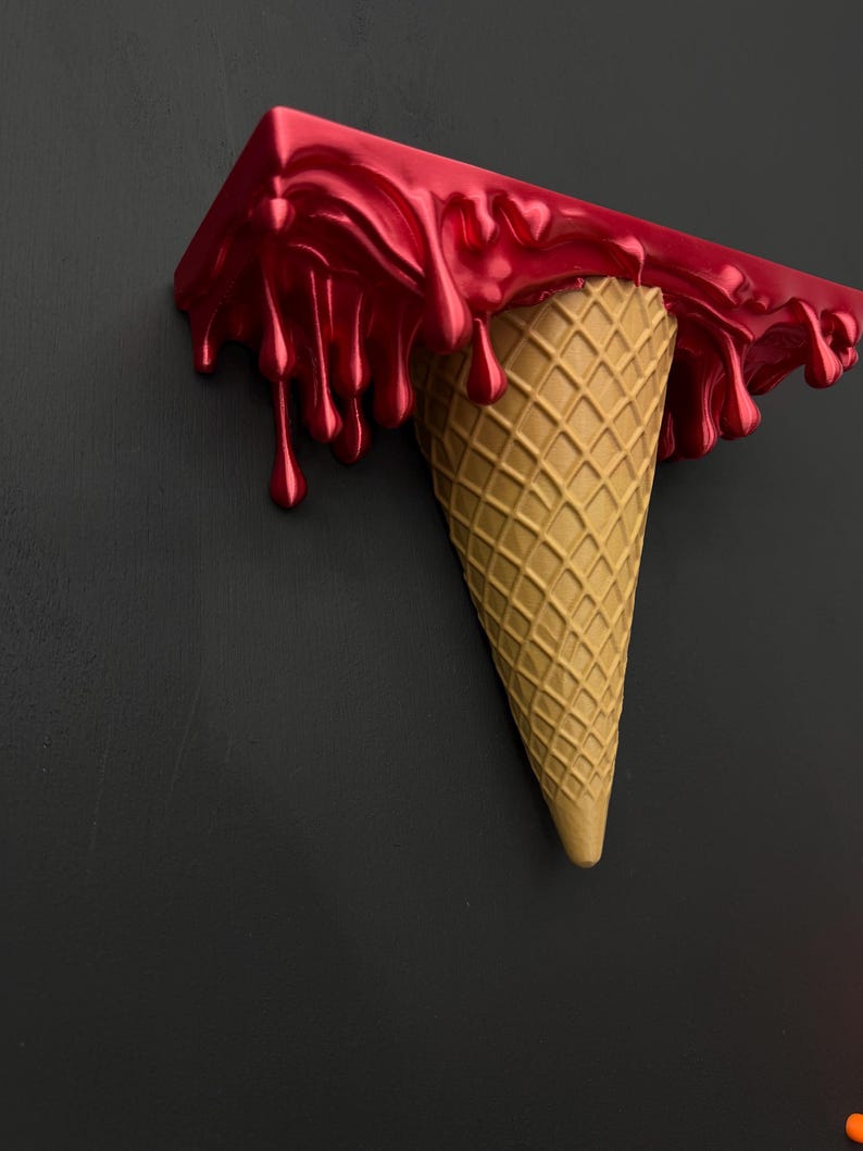 May include: A wall-mounted shelf shaped like an upside-down ice cream cone. The top section features a glossy, metallic red finish with realistic drips cascading down the sides, while the bottom is a textured, tan-colored waffle cone.
