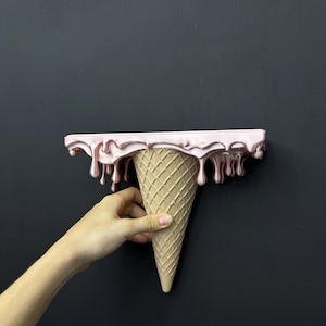 May include: A wall-mounted shelf shaped like an upside-down ice cream cone. The top flat surface features a glossy, metallic light pink finish with realistic drips cascading down the sides, while the base is a textured, tan waffle cone.