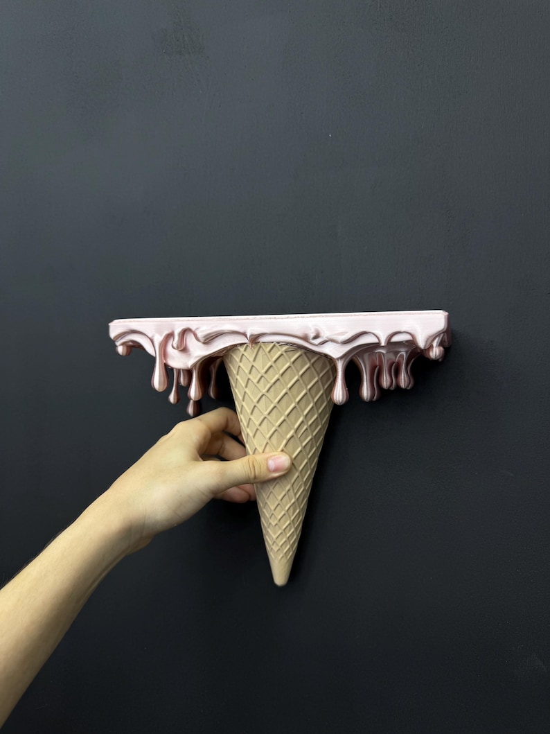 May include: A wall-mounted shelf shaped like an upside-down ice cream cone. The top flat surface features a glossy, metallic light pink finish with realistic drips cascading down the sides, while the base is a textured, tan waffle cone.