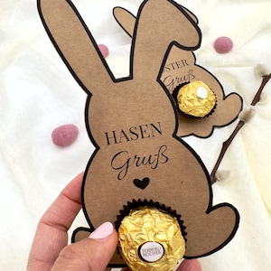 Op de afbeelding: Bruine paashaas cadeaulabels van karton met zwarte contouren, met goud omwikkelde chocolade. De labels hebben de woorden "HASEN Gruß" erop gedrukt. Roze decoratieve ballen en katjes takken zijn op de achtergrond.