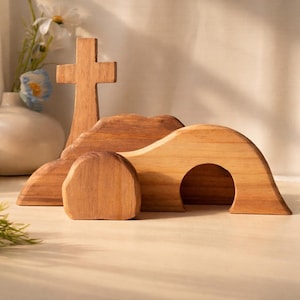 May include: Wooden Easter scene featuring a cross, a tomb, and a rolling stone. The cross stands upright, while the tomb has an arched opening. The rolling stone is a rounded shape. The scene is made of light brown wood.