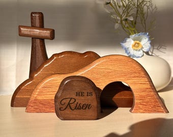 May include: Wooden Easter scene featuring a cross, rolling hills, and an open tomb. The tomb has the words "He Is Risen" engraved on it. The scene is made of various shades of brown wood. A vase of flowers is in the background.