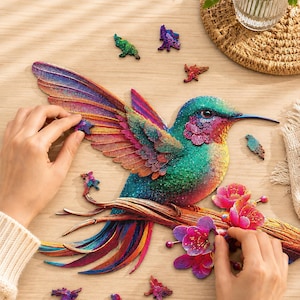 Könnte beinhalten: Ein farbenfrohes Holzpuzzle in Form eines Kolibris, der auf einem blühenden Ast sitzt. Der Vogel zeigt schillernde Töne in Türkis, Blau, Violett und Orange. Mehrere lose, individuell geformte Puzzleteile sind um das Hauptmotiv herum verstreut.