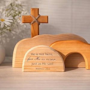 Könnte beinhalten: Osterszene aus Holz mit einem Kreuz, einem gebogenen Bogen und einer Holzstruktur mit dem Text "He is not here, for He is risen, just as He said Matthew 28:6". Die Szene ist aus hellem Holz gefertigt.