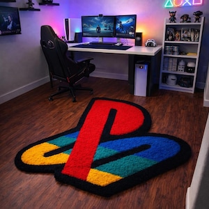 Könnte beinhalten: Ein farbenfroher Teppich in Form des PlayStation-Logos mit roten, blauen, gelben und grünen Abschnitten, die schwarz umrandet sind. Der Teppich liegt auf einem Holzboden in einem Spielzimmer, mit einem Schreibtisch, Computermonitoren und Regalen.