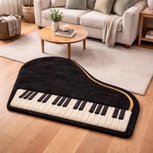 Puede incluir: Una alfombra negra con forma de piano con teclas blancas y negras y un borde dorado. La alfombra está colocada sobre un suelo de madera en un salón, con un sofá y una mesa de centro al fondo.