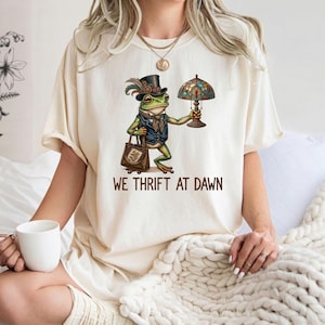 May include: A cream-colored t-shirt featuring a graphic of a green frog wearing a top hat and vest, holding a stained-glass lamp and a brown handbag. Below the frog, the text reads WE THRIFT AT DAWN in brown capital letters.