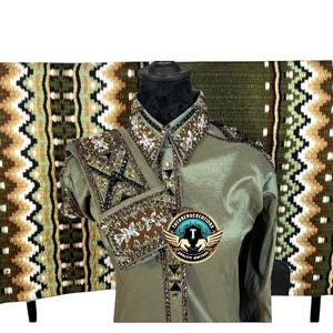Op de afbeelding: Een saliegroen shirt met lange mouwen en gedetailleerd bruin en zilverkleurig kraalwerk op de kraag en manchetten, getoond op een paspop voor een rechthoekig zadeldek met een herhalend geometrisch patroon in olijf, bruin, wit en zwart.