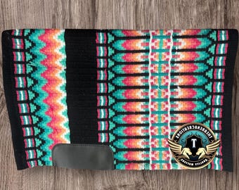 Pode incluir: Uma almofada de sela de cavalo colorida com um padrão geométrico em verde-azulado, coral e branco sobre um fundo preto. A almofada tem um remendo de couro preto e um logotipo. Projetada para uso equestre.
