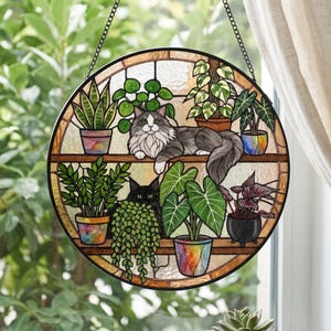 May include: A circular stained glass suncatcher featuring two cats resting on shelves among various potted plants. The top shelf holds a long-haired gray and white cat, while the bottom shelf features a black cat surrounded by green foliage.