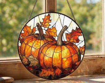May include: A circular stained glass suncatcher featuring two orange pumpkins surrounded by vibrant autumn leaves in shades of red, yellow, and orange. The piece is framed in black metal and hangs from a thin metal chain against a window.